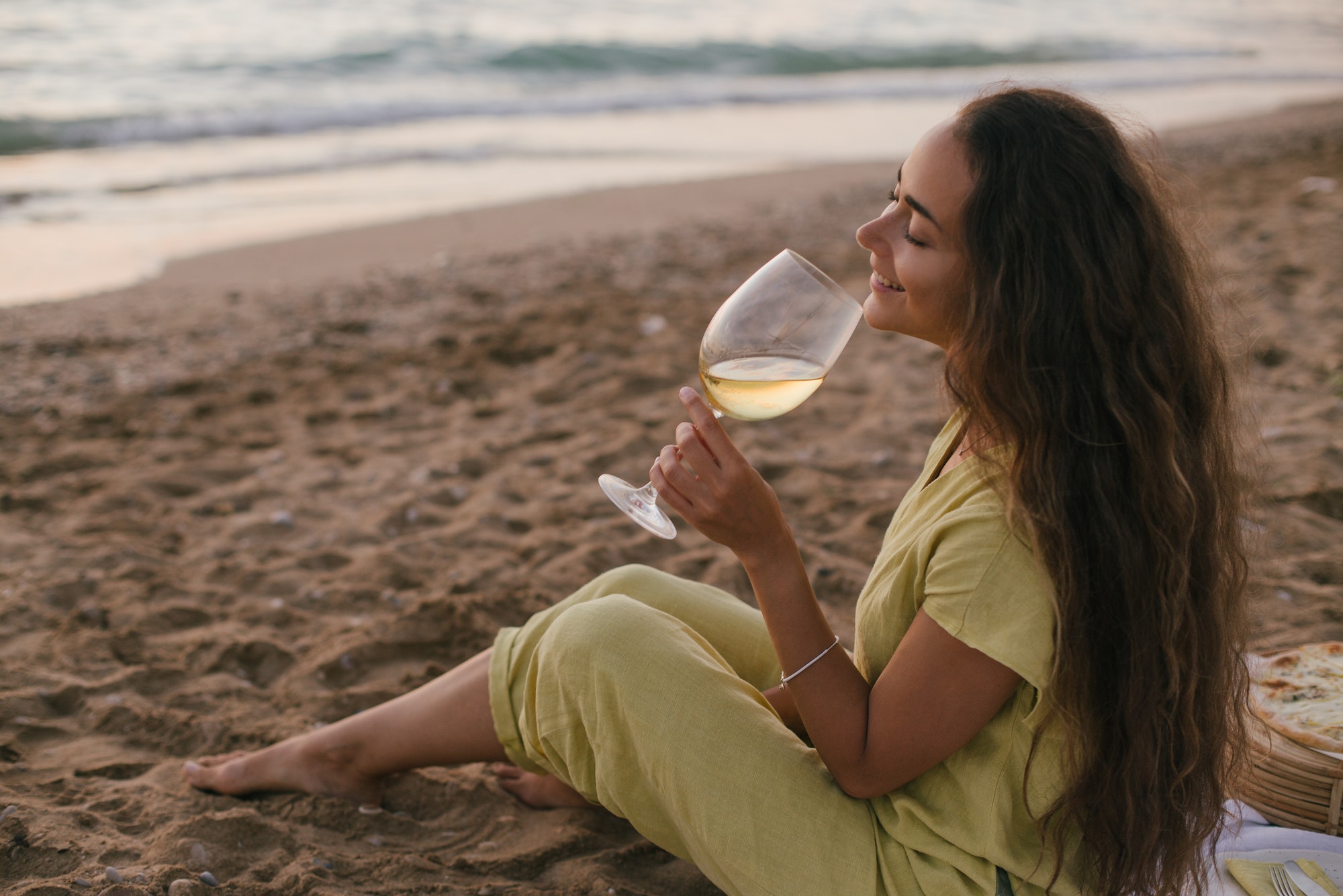 Young beautiful woman enjoying picnic with pizza and glass of wine sitting on a beach at sunset.