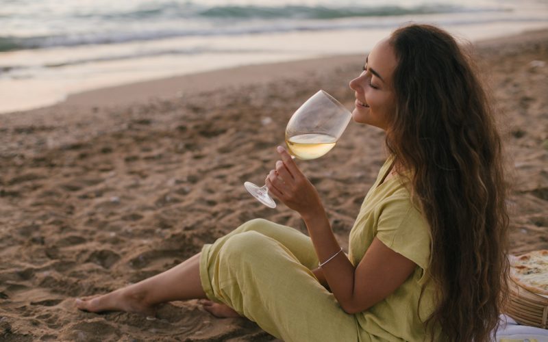 Young beautiful woman enjoying picnic with pizza and glass of wine sitting on a beach at sunset.